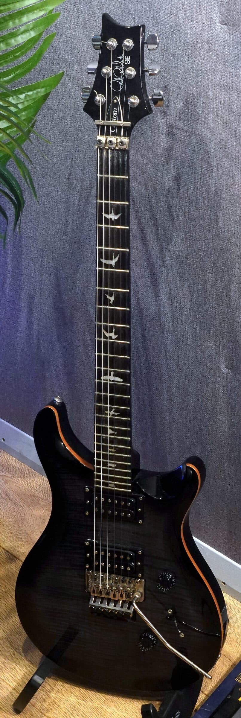 A PRS electric guitar on a stand against the fabric wall, a GT-3 pedalboard at its feet